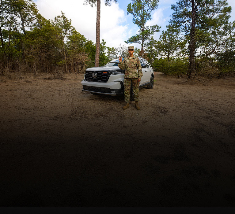 2025 Honda Pilot TrailSport in Platinum White Pear front on a dirt path in the woods with a soldier (Full-Size SUV).