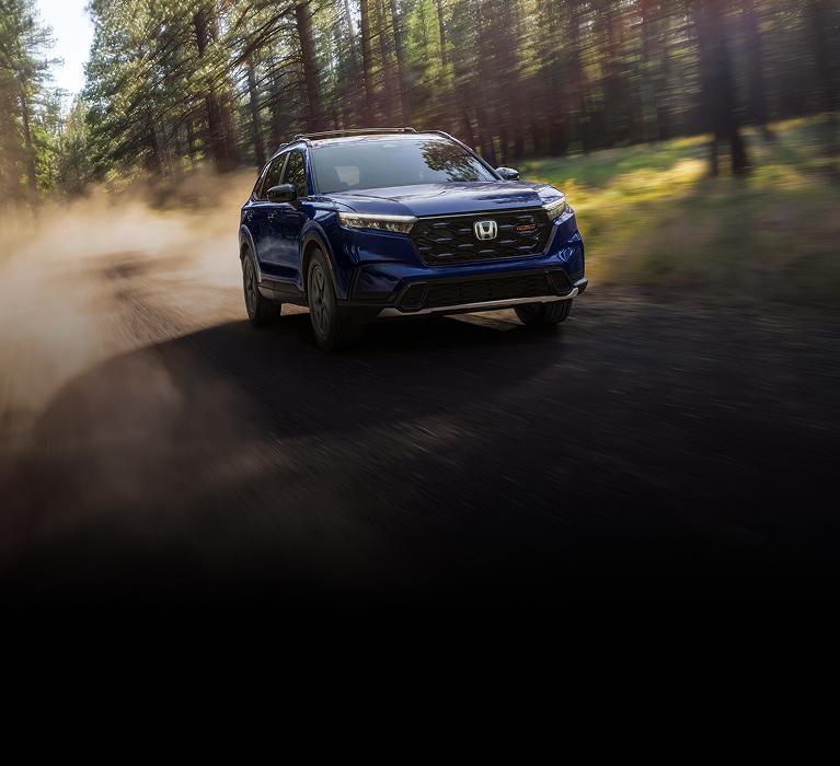 2026 Honda CR-V Trailsport in Canyon River Blue front driving on a dirt road through a forest (Compact Crossover SUV).