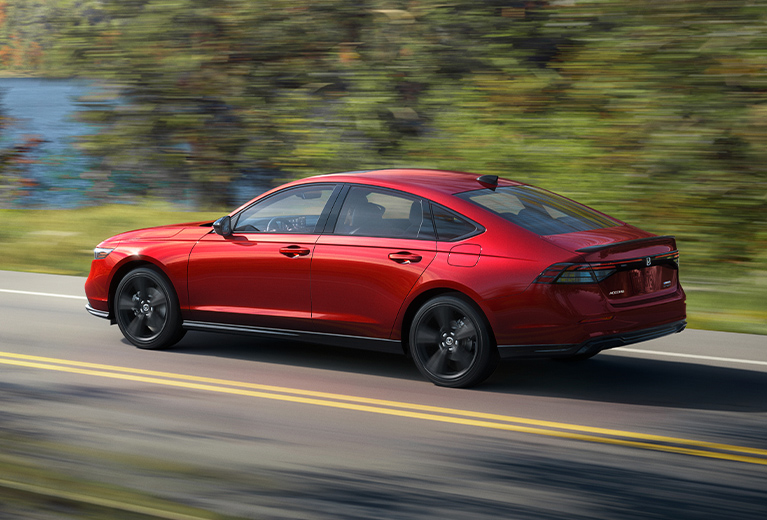 2023 Honda Accord Sport L in Radiant red Metallic profile driving on a scenic road past trees and water (Mid-Size Sedan).